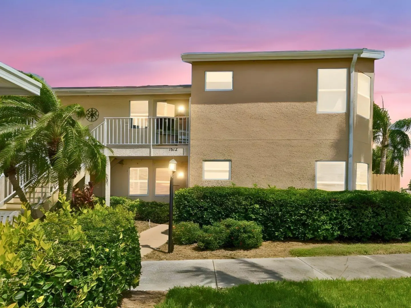 Two-story tan apartment building with white trim, balcony, and palm trees against a pink and purple sunset sky.