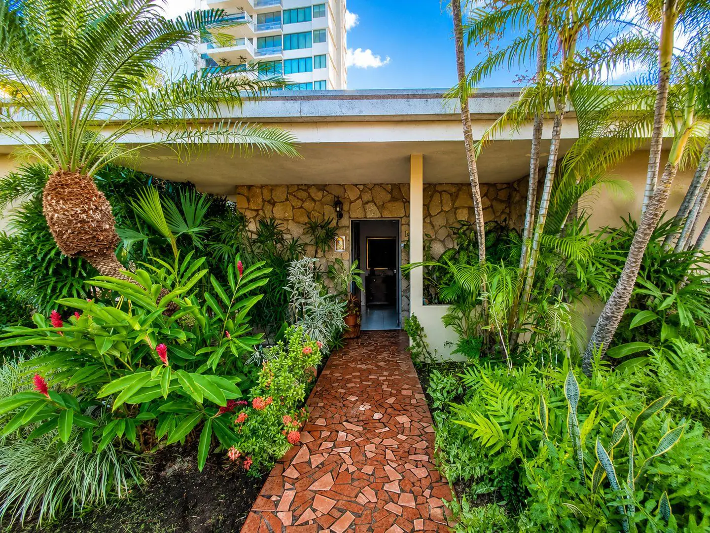Home exterior with a stone facade, lush tropical plants, and a red brick walkway leading to a dark front door. A tall building is in the background.