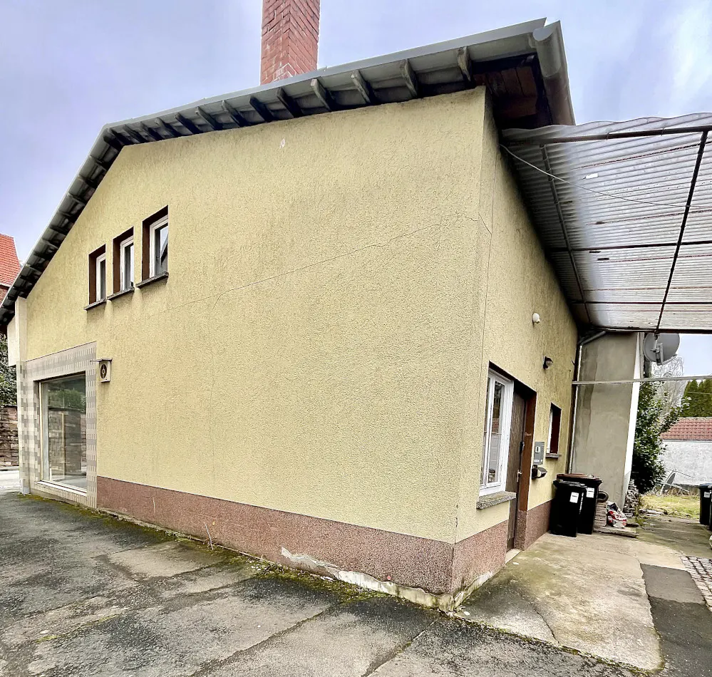 Exterior view of a yellow stucco building with a brown base, small windows, and a chimney. A covered carport is attached to the side.