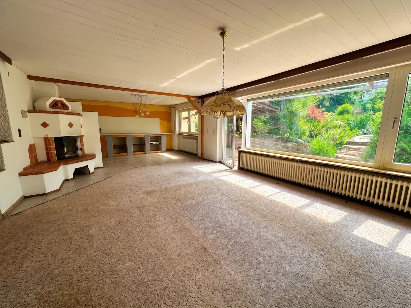 A bright, empty living room with a fireplace, open kitchen, and large window overlooking a lush garden.