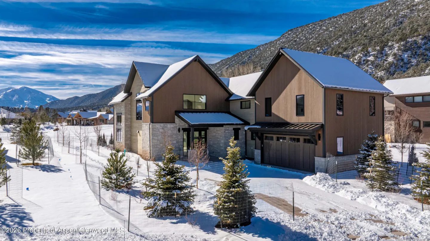 Exterior view of a modern two-story home with stone and brown siding, snow-covered roof, driveway, and yard. Mountains in the background.