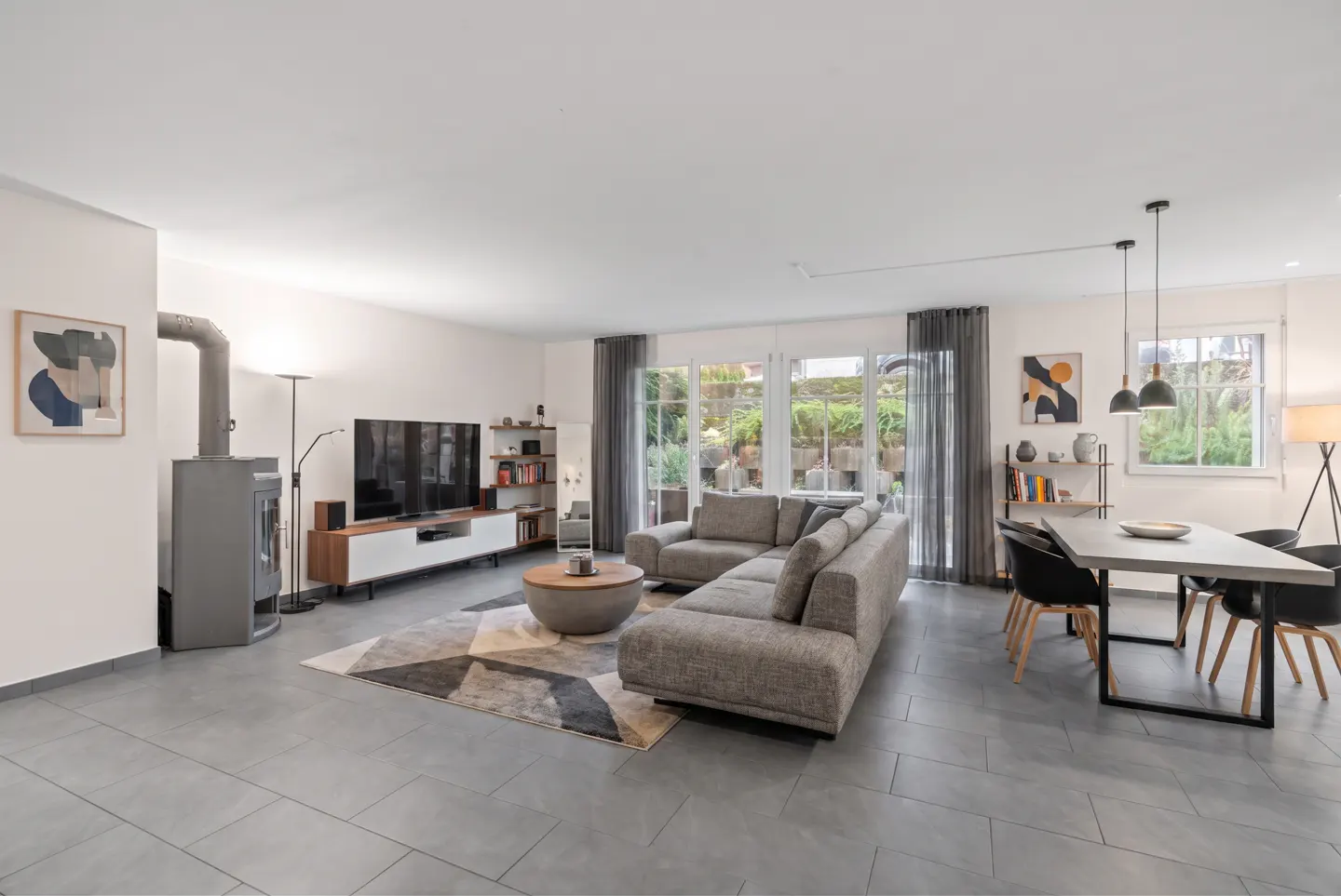 Open-concept living space with gray tile flooring, a gray sectional sofa, a dining table, and a fireplace. Large windows offer a view of the outdoors.