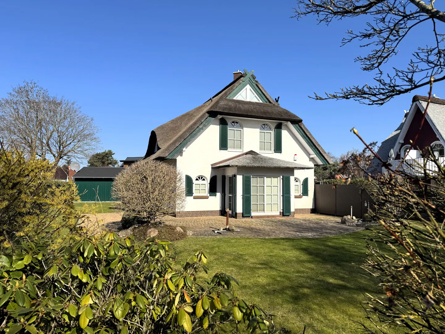 White house with a thatched roof and green shutters, surrounded by a green lawn and bushes under a blue sky.