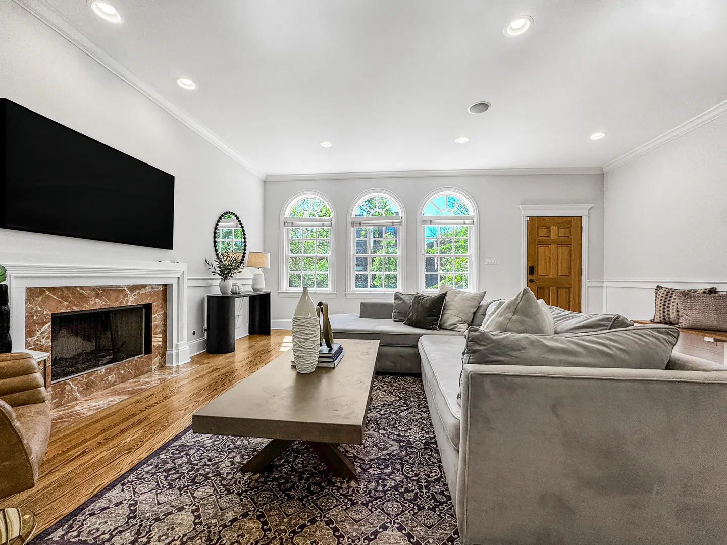 Bright living room with a gray sectional sofa, marble fireplace, and three arched windows. A patterned rug covers the hardwood floor.