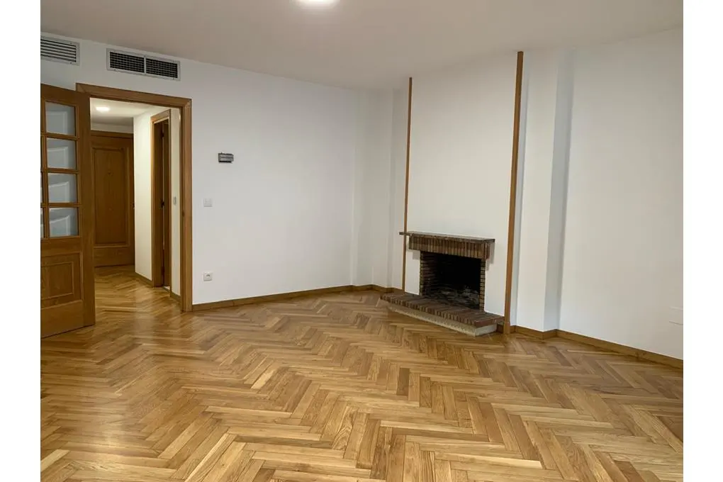 Bright, empty living room with herringbone wood floors, white walls, and a brick fireplace. A doorway leads to another room.