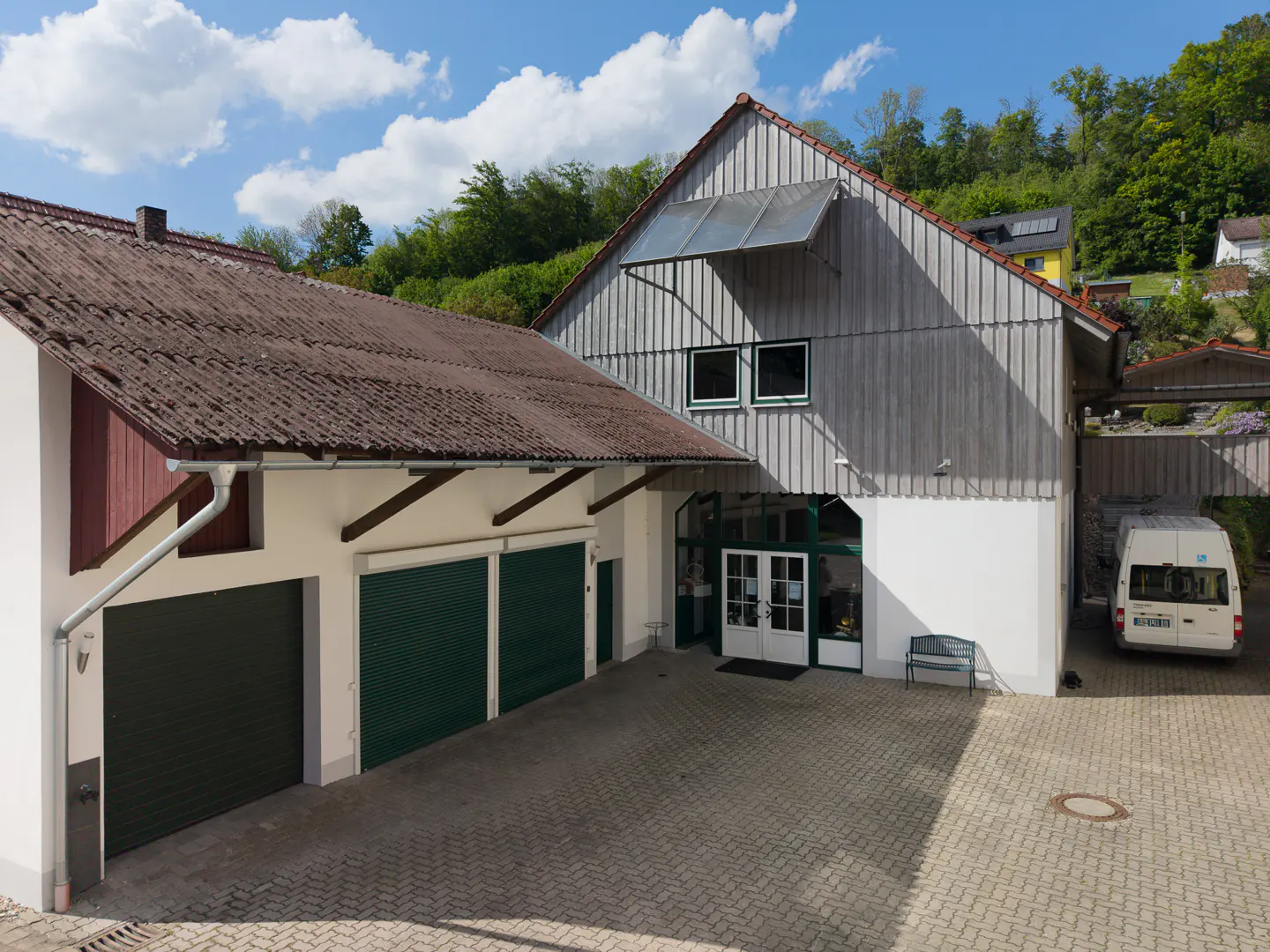 Exterior view of a two-story building with a gray wood facade, white trim, and a brown tile roof. Green garage doors and a white van are visible.