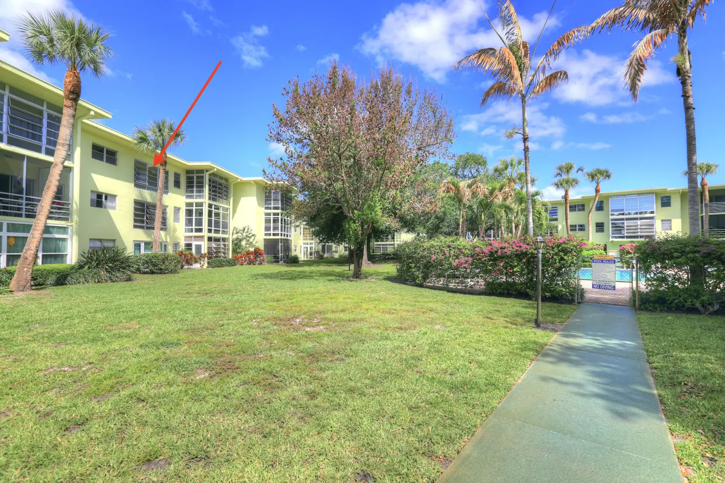 Exterior view of a light yellow apartment building with palm trees and a green lawn under a blue sky.