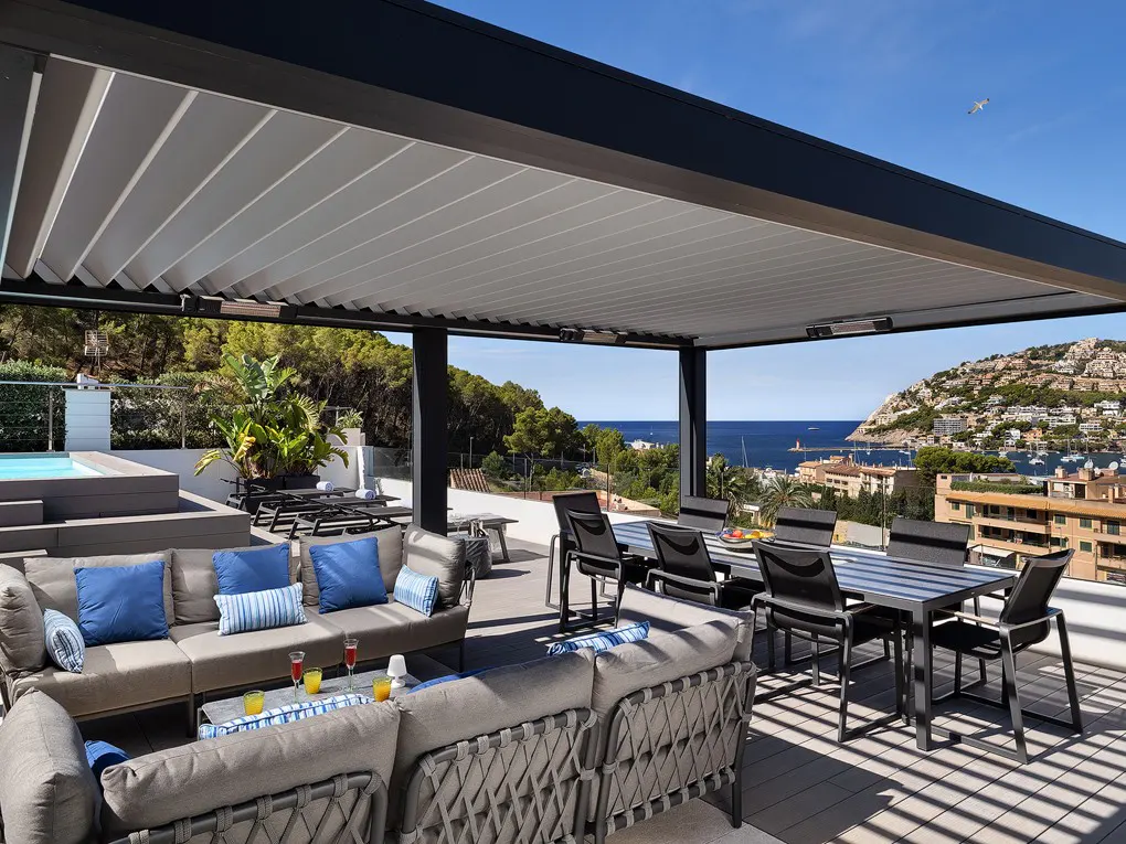 Luxury rooftop patio with gray furniture, blue pillows, and a dining table under a modern pergola overlooking the ocean and hillside.