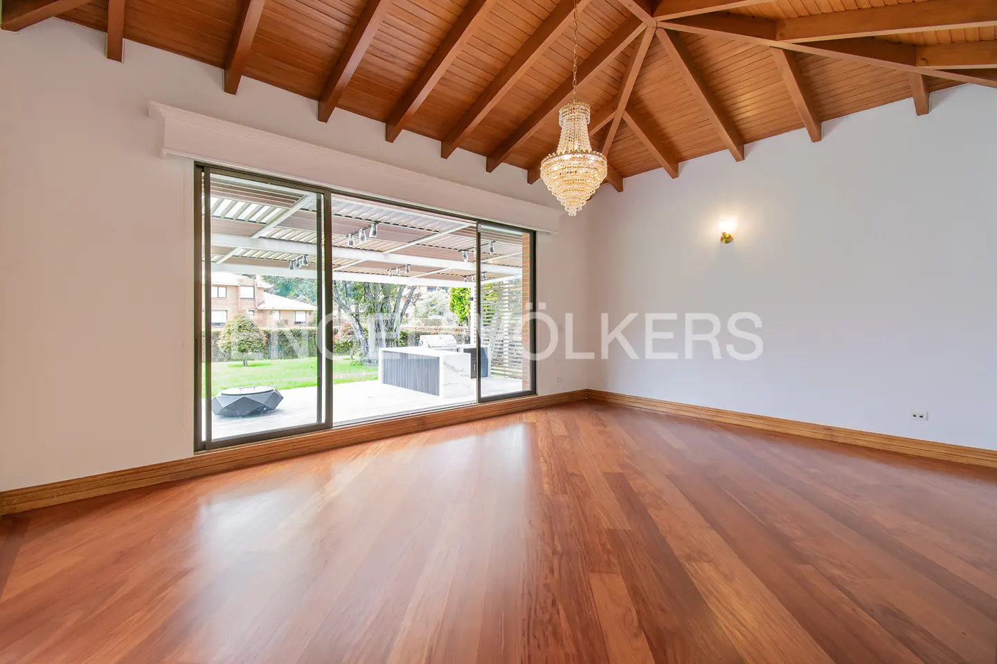 An empty room with hardwood floors, a wood-paneled ceiling, and a crystal chandelier. Sliding glass doors lead to a patio and green lawn.