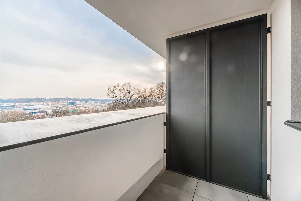 Balcony view with a white railing, gray tile floor, and black sliding doors. A cityscape is visible in the distance under a cloudy sky.