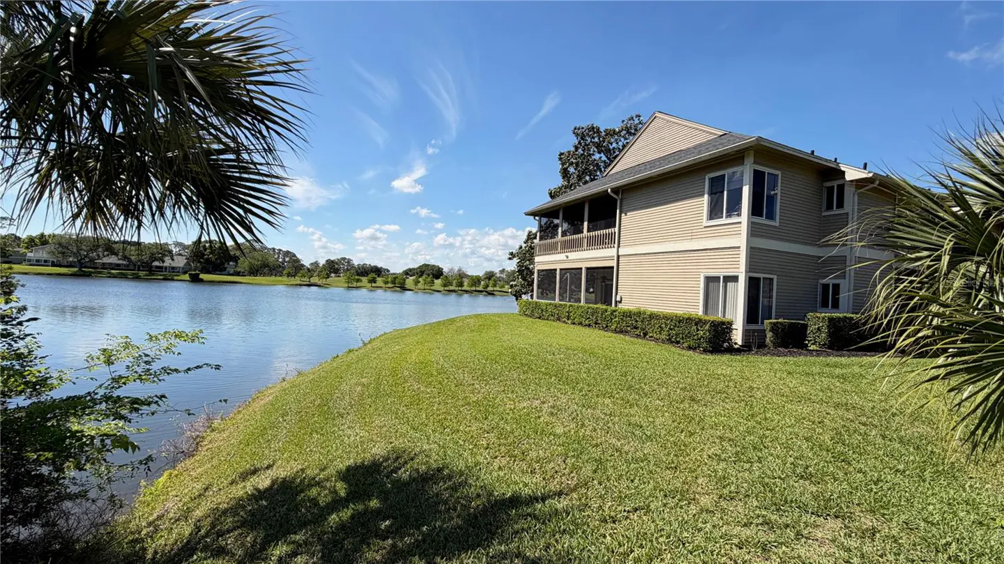 Two-story beige house with a screened porch overlooking a lake, green grass, and palm trees under a blue sky.