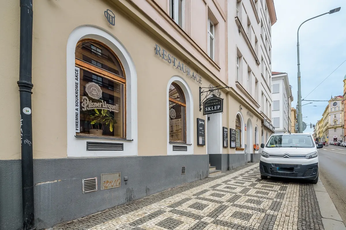Street view of Restaurace Sklep, a Pilsner Urquell restaurant in Prague, with a white van parked on the patterned sidewalk.