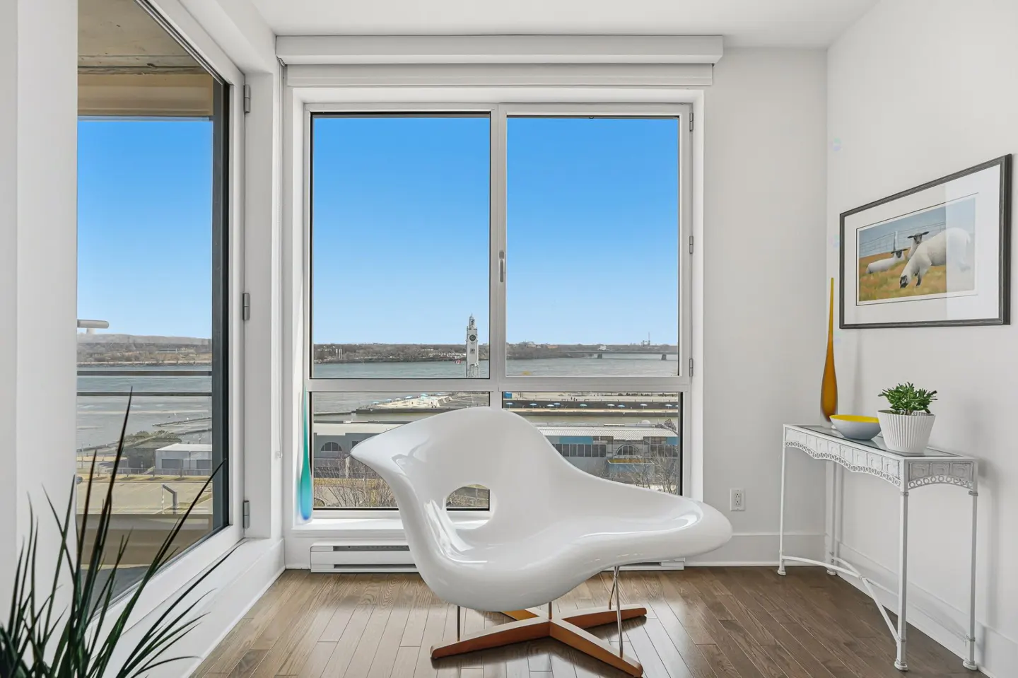 Bright room with a white chair, table, and large window showing a city view. The floor is wood and there is art on the wall.