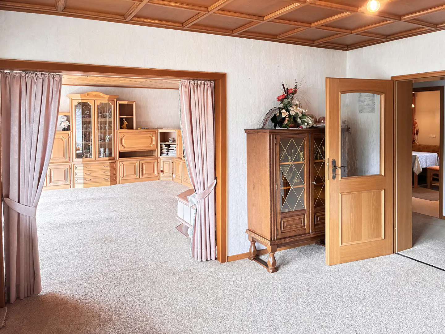 A living room with beige carpet, wood ceiling, and a large wooden cabinet. Pink curtains frame an opening to another room with a built-in wooden cabinet.