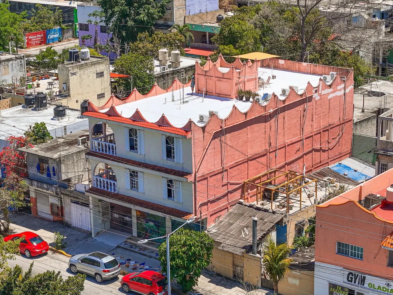 Aerial view of a three-story building with a unique crown-shaped roof, painted in pink and white, surrounded by other buildings and trees.
