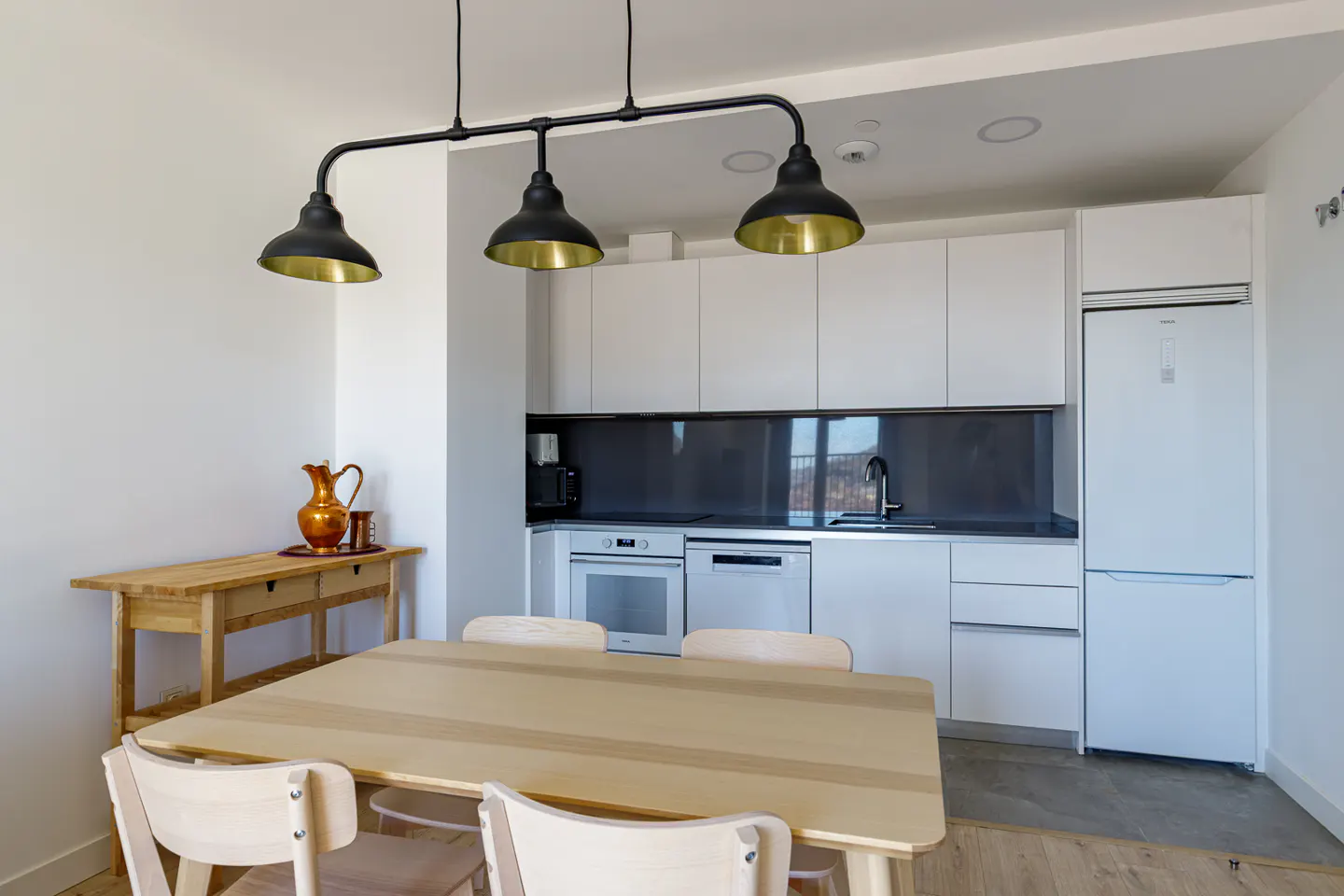 Bright kitchen with white cabinets, black backsplash, and stainless steel appliances. A wooden table and chairs sit under a black and gold pendant light.