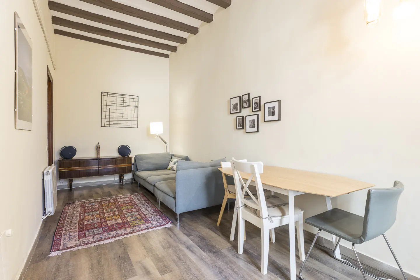 Living room with a gray sofa, wooden table with chairs, and a vintage cabinet on a patterned rug. Beamed ceiling.