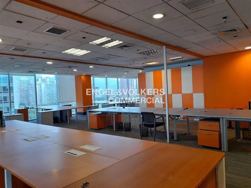 Bright, modern office space with orange accent walls, large windows, and multiple desks. Engel & Volkers Commercial logo visible.