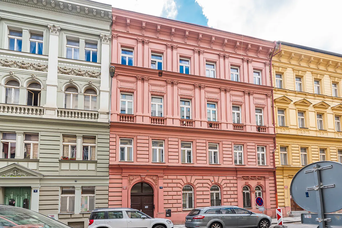 A row of historic European buildings in pastel green, pink, and yellow, with a silver car parked on the street in front.