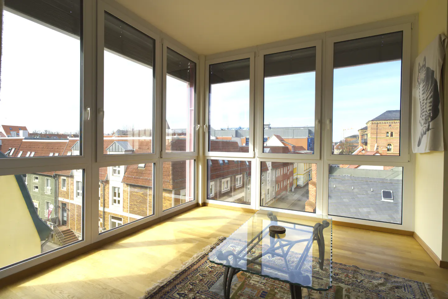 Bright sunroom with a glass table on a rug, surrounded by large windows showing a cityscape with red-roofed buildings.