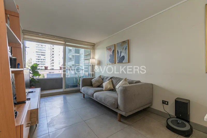 Living room with a gray sofa, pillows, art, and a balcony view of city buildings. A robot vacuum is near a black charging station.
