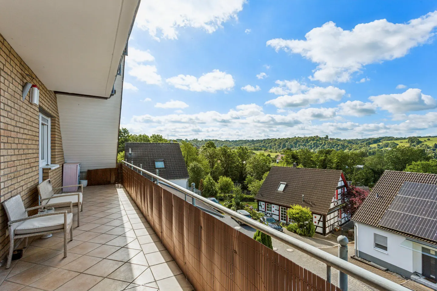 Balcony with tiled floor and two chairs overlooking a scenic neighborhood with traditional houses and lush green hills.