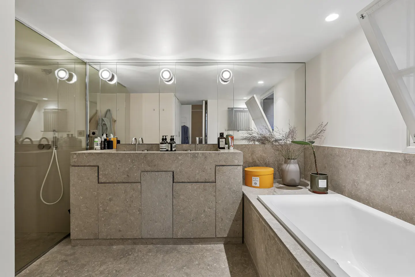 Modern bathroom with gray stone vanity, double sinks, and a large mirror with round lights. A white bathtub is on the right.