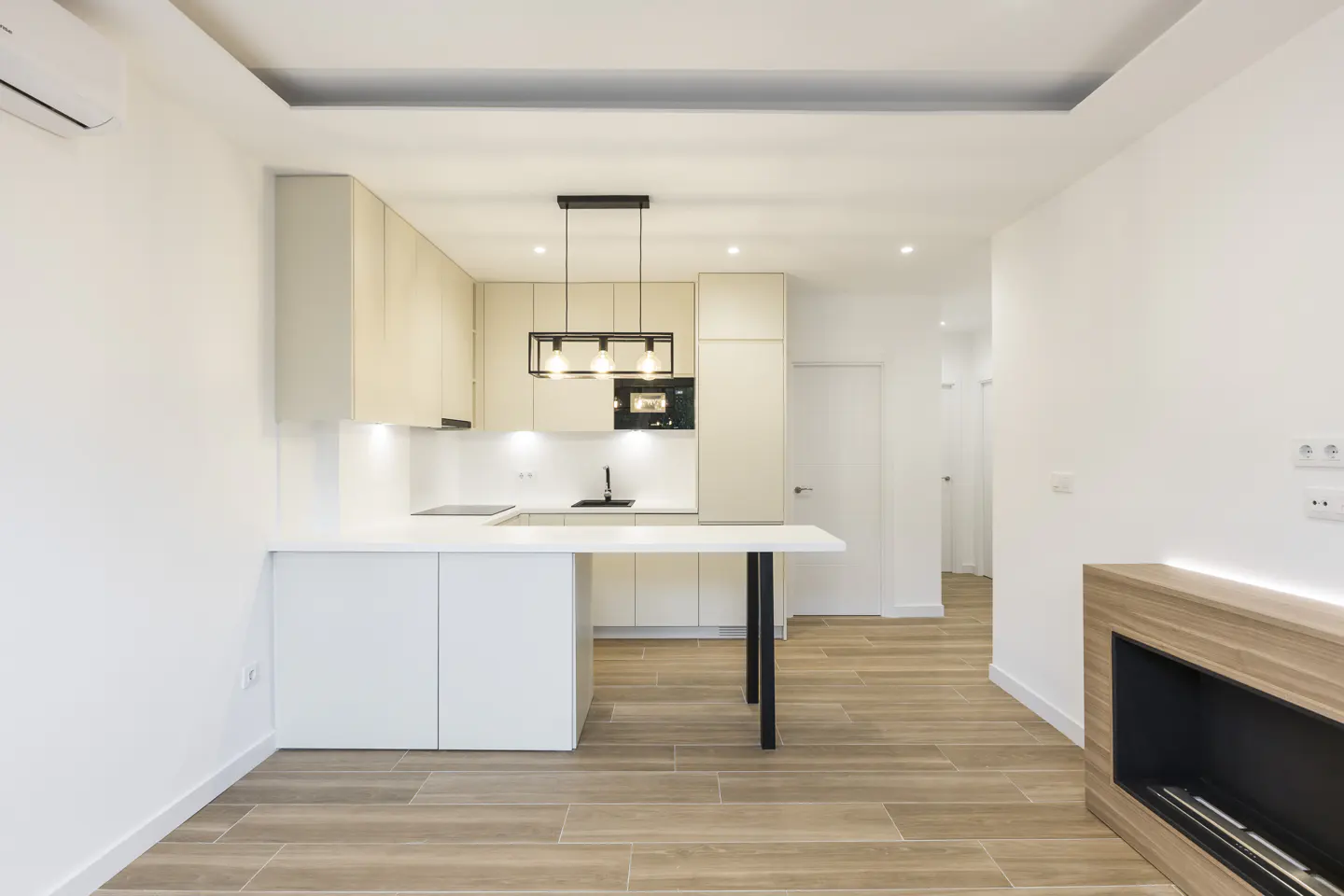 Bright, modern kitchen with white cabinets, light wood accents, and a black pendant light. Wood-look tile floors lead to a fireplace.