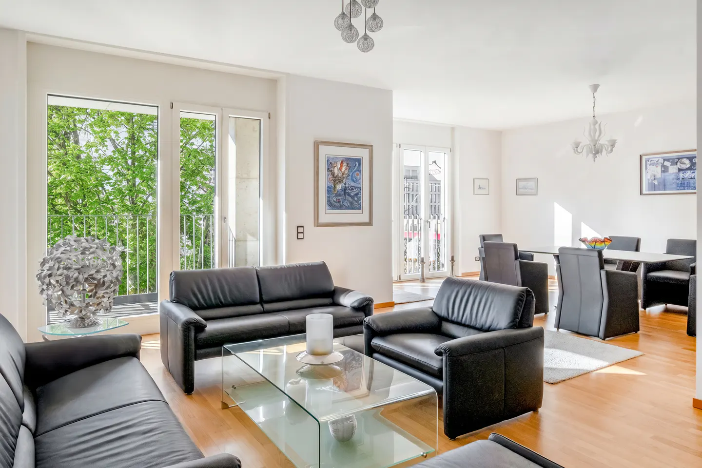 Bright living room with black leather sofas, glass coffee table, and dining area with gray chairs. Balcony with green trees visible.