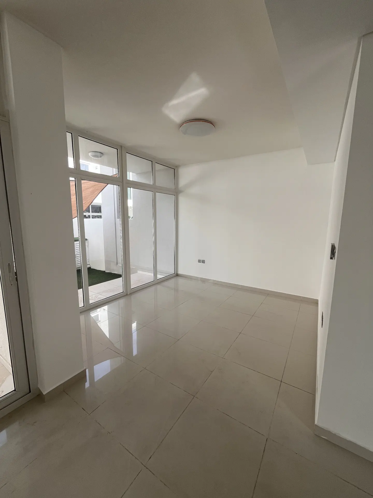 Bright, empty room with white walls and tile floor. Large glass doors lead to a patio with artificial grass. Ceiling light fixture visible.