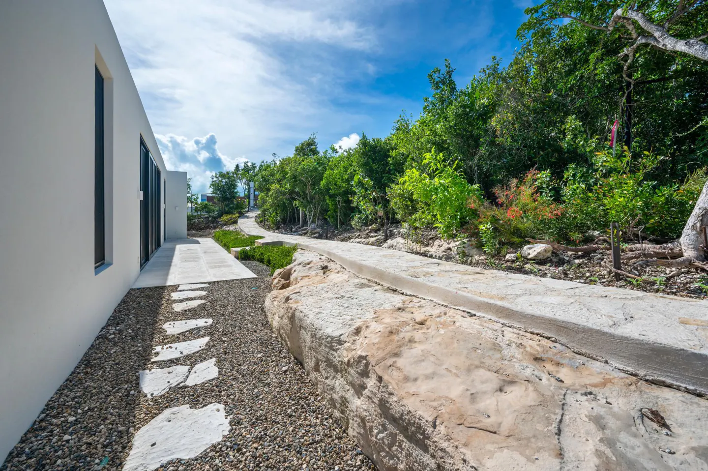 Exterior view of a modern white house with a stone pathway leading to lush green trees under a blue sky.