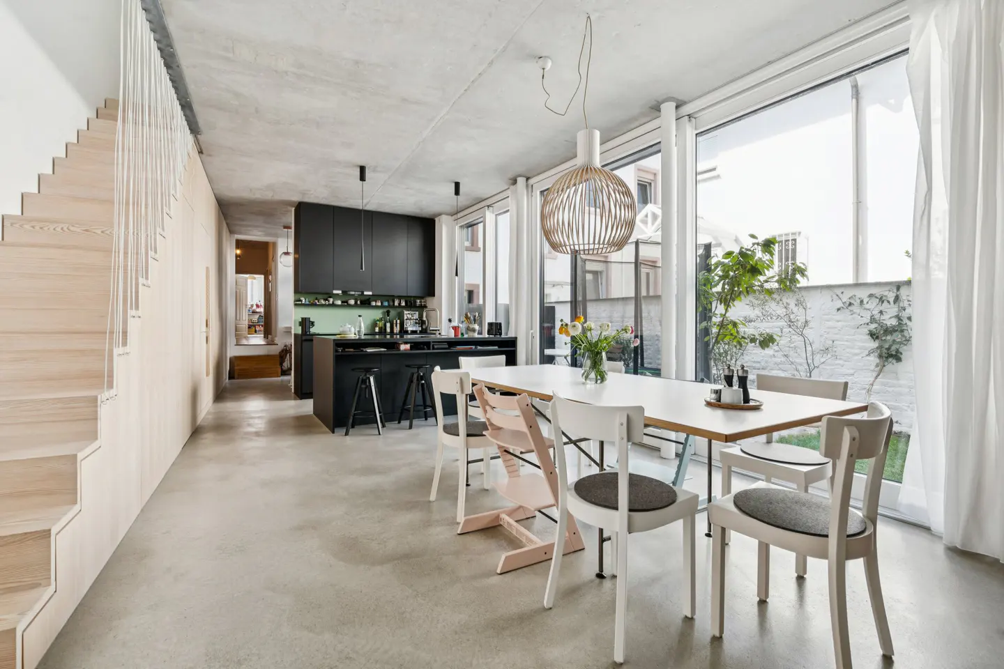 Bright, modern interior with a dining table, white chairs, and a black kitchen island. Large windows offer a view of a garden. A wooden staircase is on the left.
