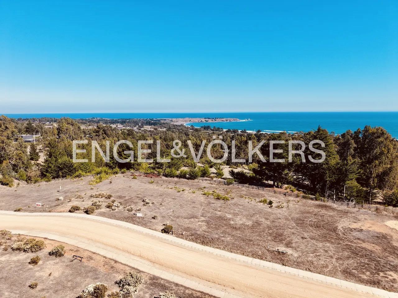 Scenic view of land for sale with ocean backdrop under a clear blue sky. Engel & Völkers logo overlayed on trees.