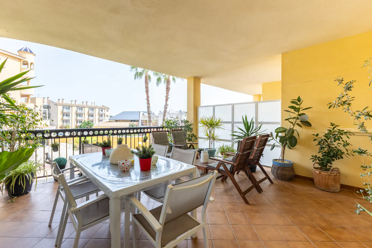 Outdoor patio with tile floor, yellow walls, table, chairs, and plants. Balcony overlooks buildings and palm trees.