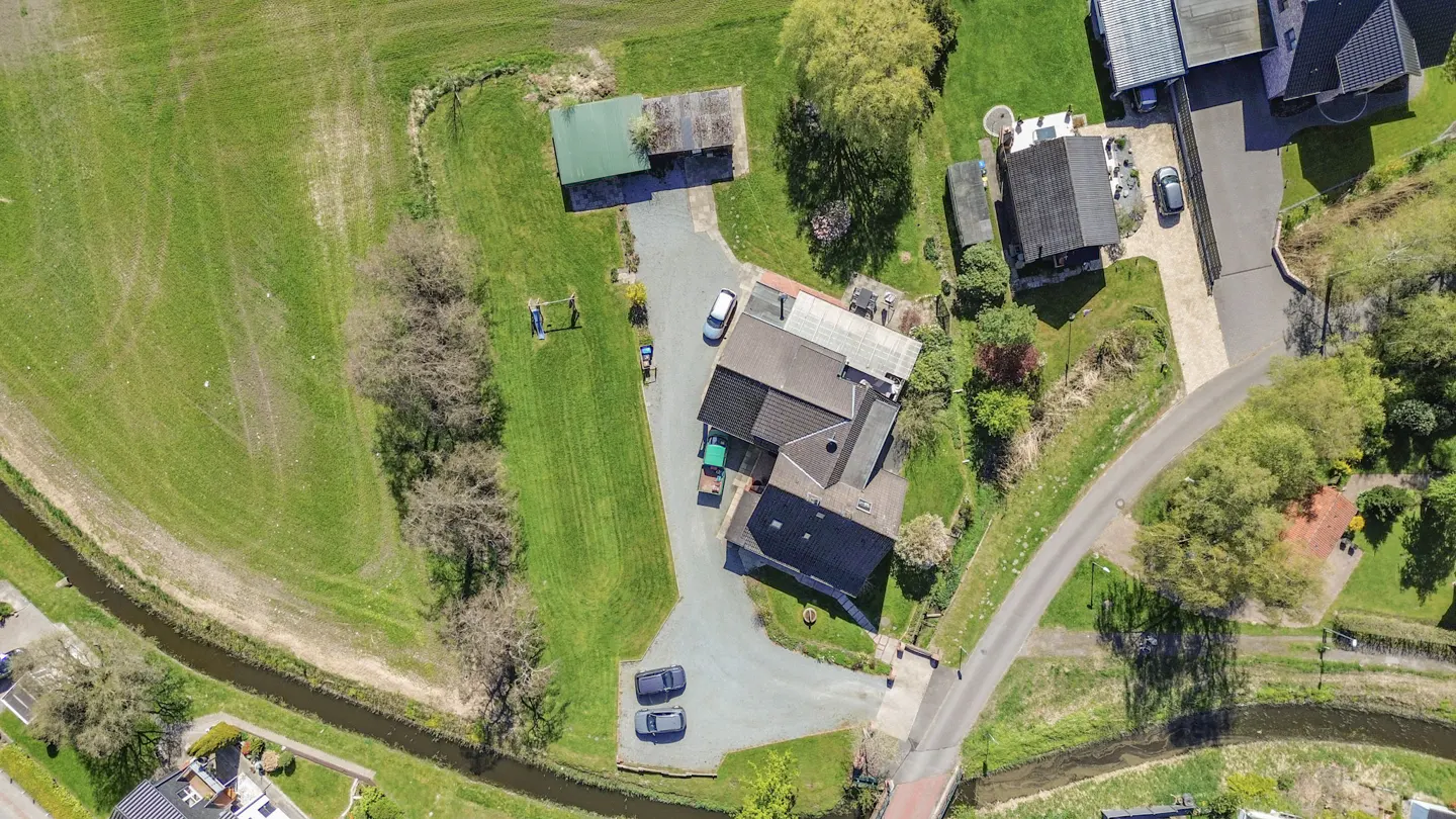 Aerial view of a house with a dark roof, surrounded by green grass, trees, and a driveway with parked cars.