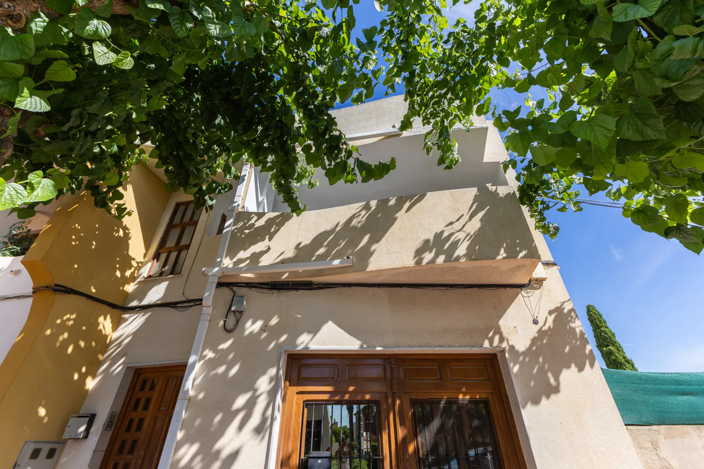 Low angle view of a two-story building with a yellow and beige facade, brown doors, and green foliage overhead against a blue sky.