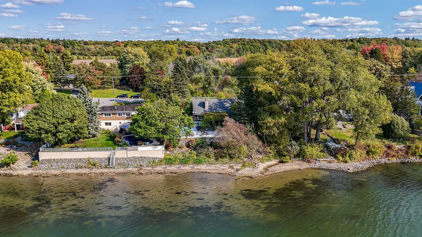 Aerial view of waterfront homes with trees in autumn colors, and clear water under a partly cloudy sky.