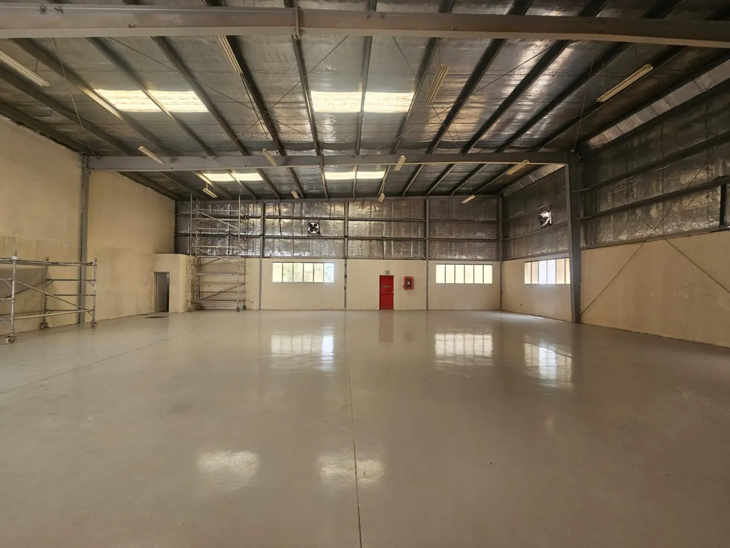 Empty warehouse interior with a gray floor, metal ceiling, and a red door on the back wall.