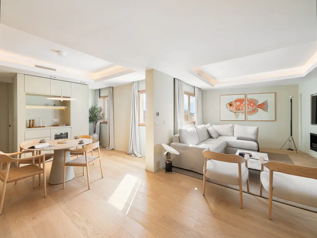 Open-concept living space with light wood floors, a dining table with chairs, and a white sectional sofa. Artwork of a fish hangs on the wall.