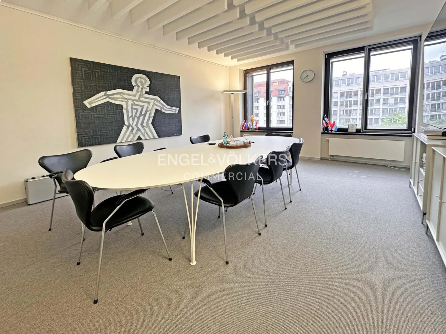 Bright office space with a white oval table, black chairs, and a modern art piece on the wall. Windows overlook city buildings.