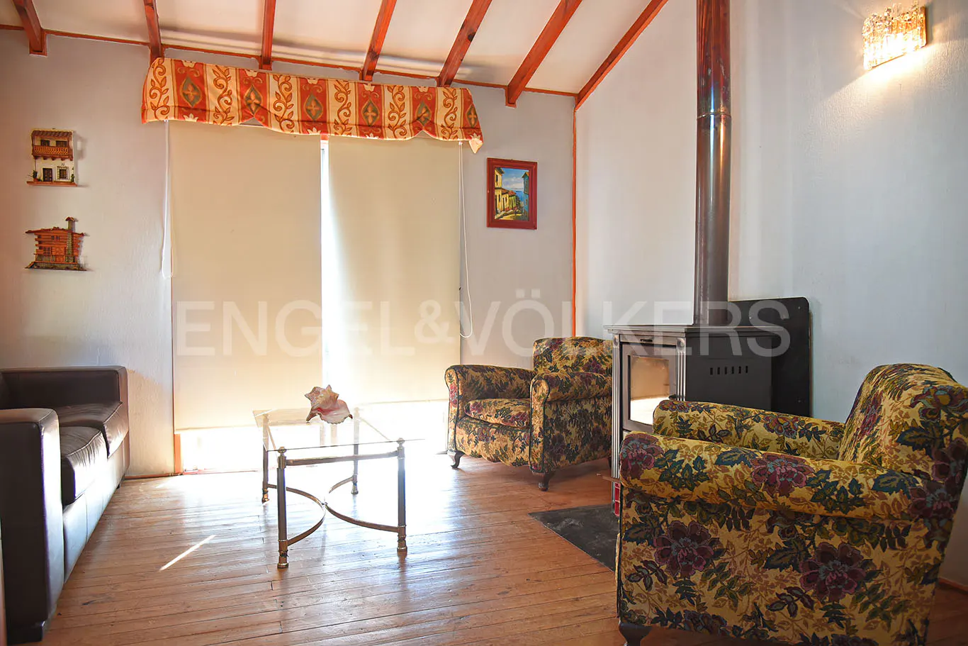 Living room with wood floors, floral armchairs, and a black fireplace. A brown sofa sits near a glass coffee table. Orange beams accent the ceiling.