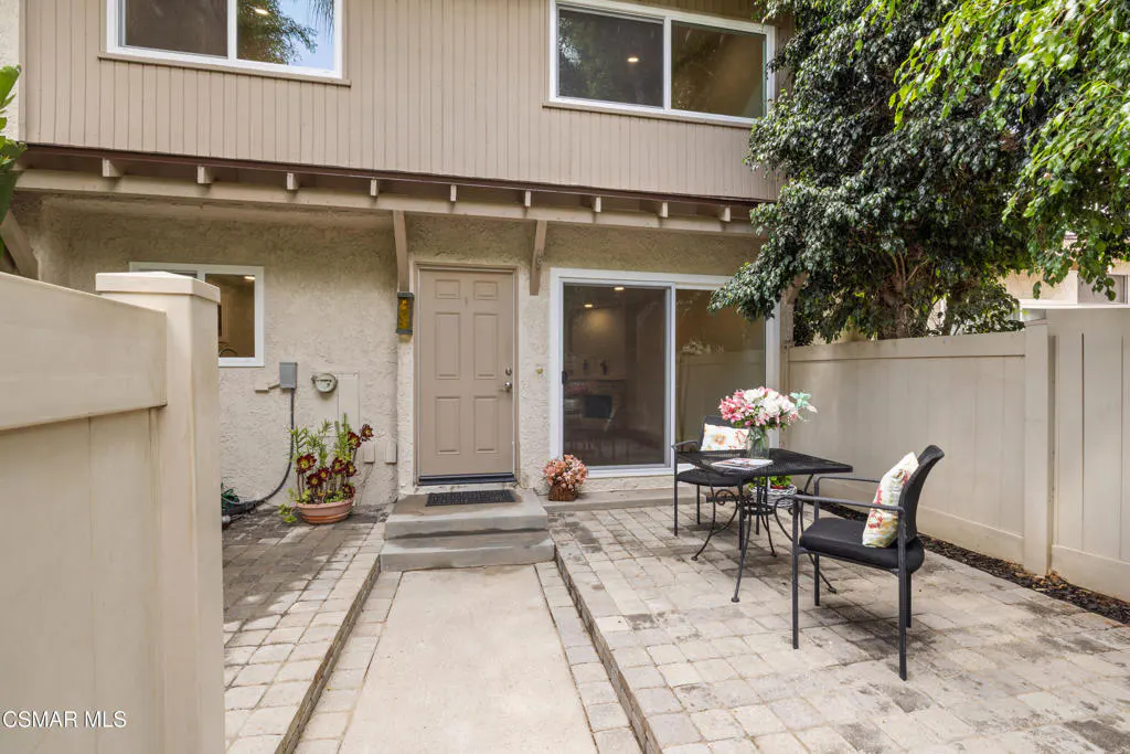 Tan townhouse exterior with a brick patio, table, chairs, and a sliding glass door. A tree is visible in the background.