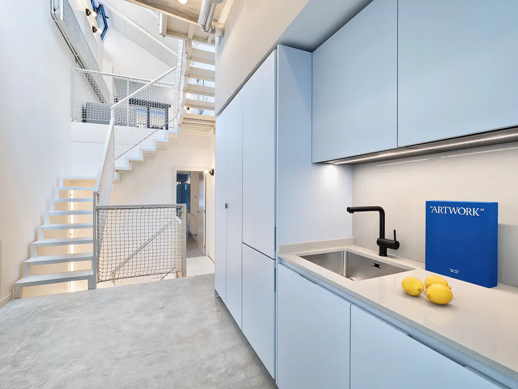Modern kitchen with light blue cabinets, black faucet, and stainless steel sink. Lemons and a blue "Artwork" book sit on the counter. White stairs lead to a loft area.