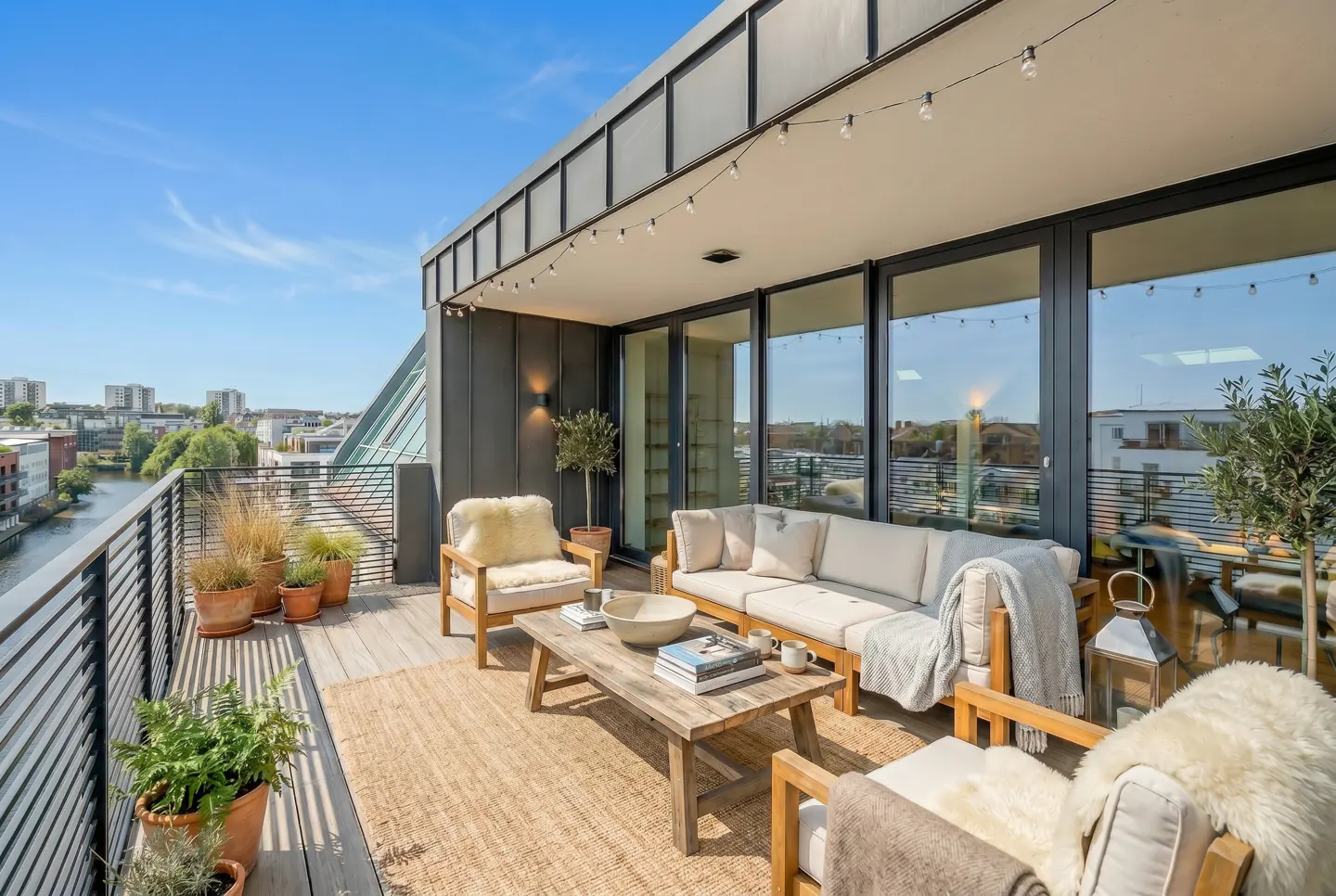 Modern rooftop terrace with outdoor sofa, armchairs, wooden coffee table, potted plants, and string lights overlooking a river.