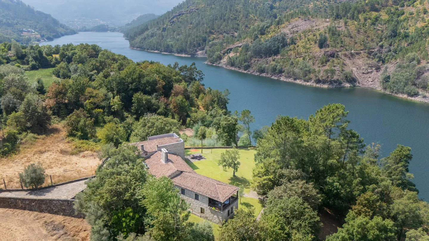 Aerial view of a stone house with a tiled roof nestled in a lush green forest beside a winding river and mountains.