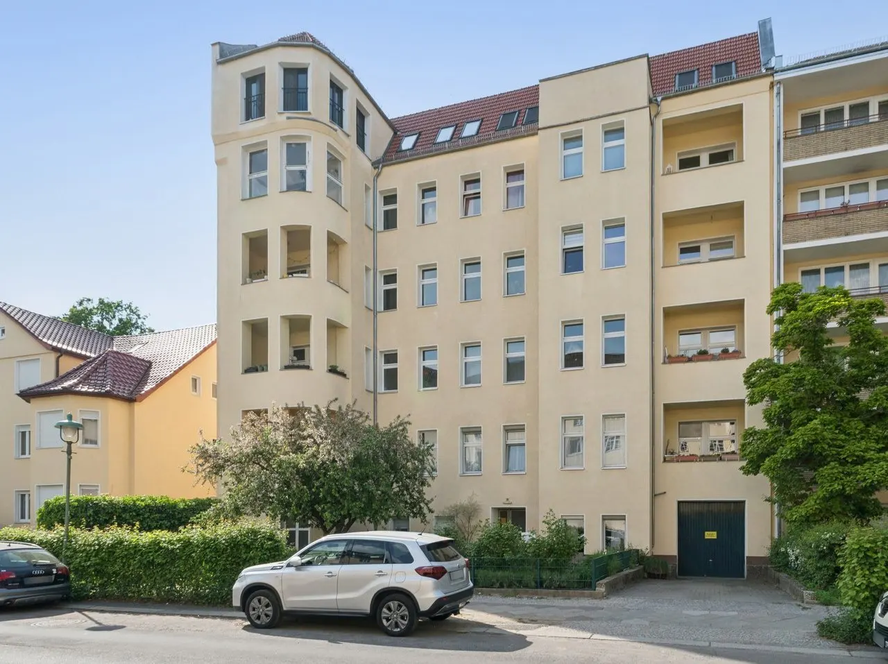 Exterior view of a multi-story, light yellow apartment building with a rounded corner, cars parked in front.