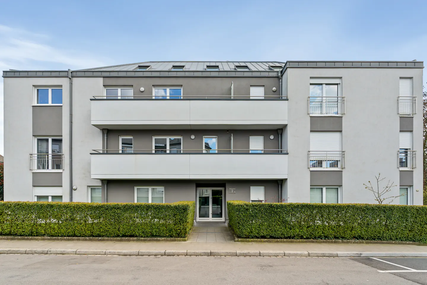 Three-story gray apartment building with balconies and a green hedge in front.