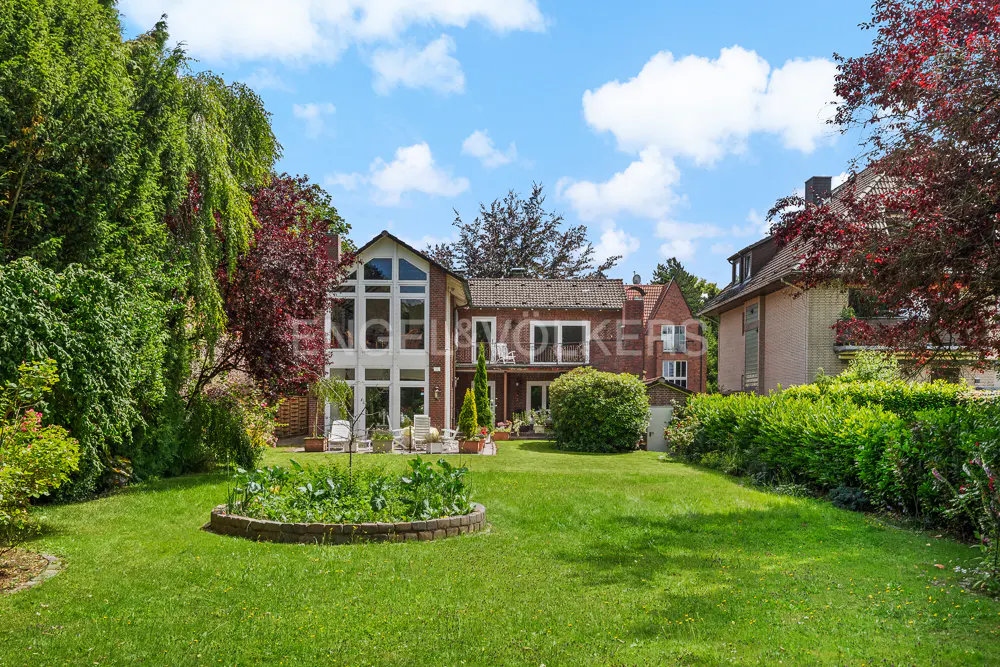 Backyard view of a brick house with a large lawn, trees, and blue sky. The house has white framed windows and a balcony.