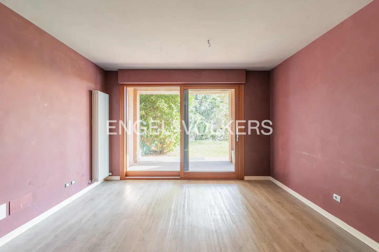 Empty room with mauve walls, light wood floors, and sliding glass doors to a green yard. Engel & Volkers logo on the glass.