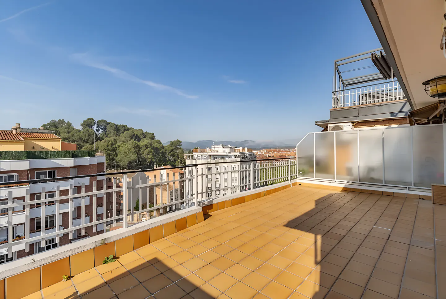 Wide, sunny terrace with yellow tile flooring and white railings overlooking a cityscape and distant mountains under a clear blue sky.